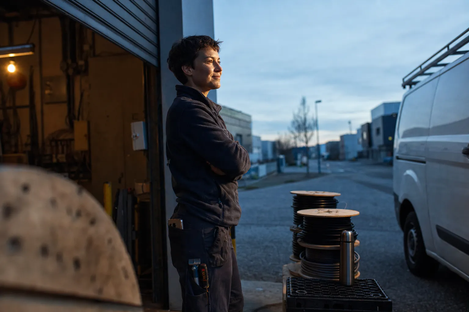 Jeune électricienne devant son atelier au petit matin, regard vers la rue